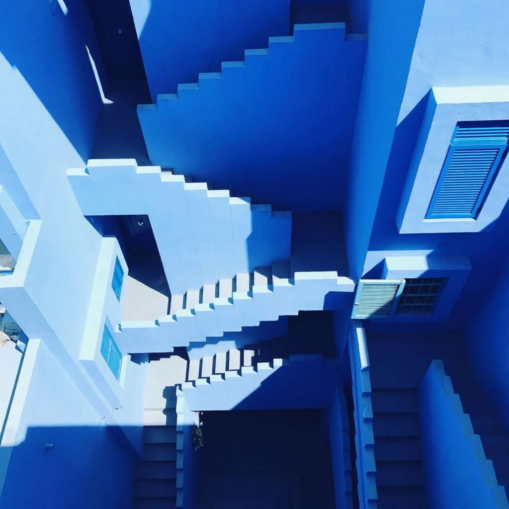 A geometric view of multiple staircases in a building with walls, doors, and windows painted in various shades of blue, designed using TouchDesigner. Shadows cast by the structures enhance the angular design.