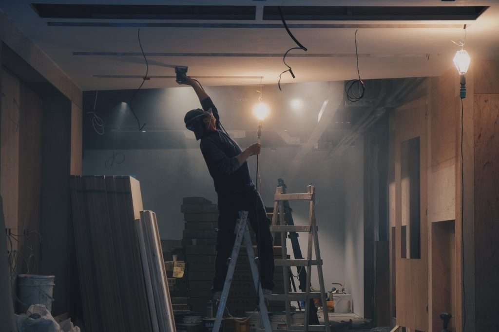 A person stands on a ladder, working on the ceiling of a partially constructed room. Building materials and tools are scattered around. The space, resembling a TouchDesigner workspace, is dimly lit by exposed lightbulbs.