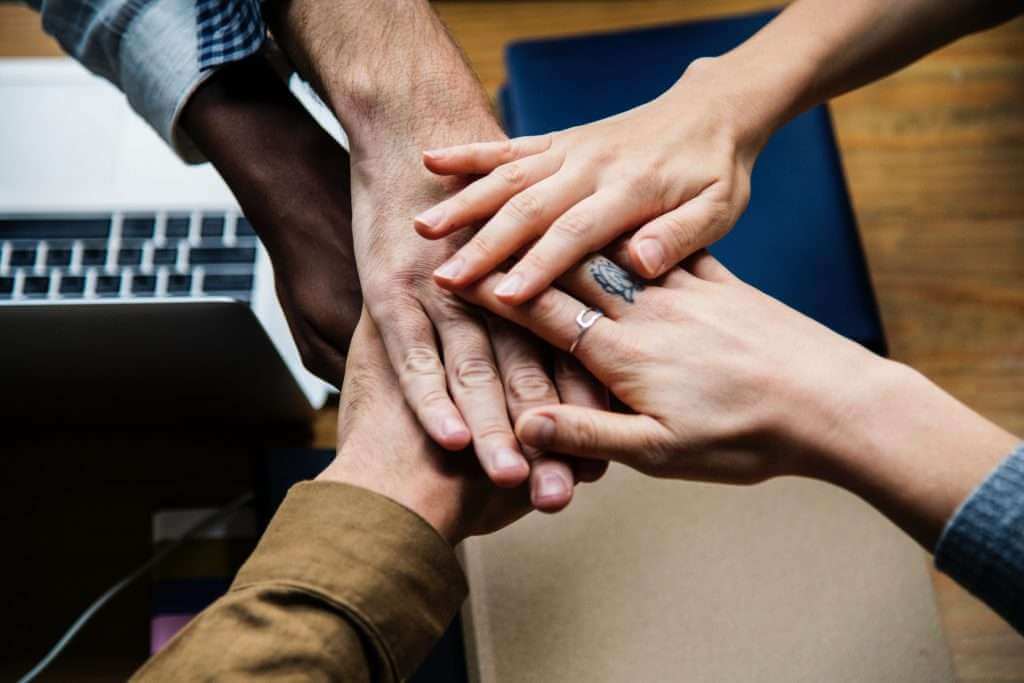 Multiple hands placed on top of each other over a table beside a laptop and notebooks, symbolizing teamwork and collaboration—much like the seamless integration achieved with TouchDesigner in creative projects.