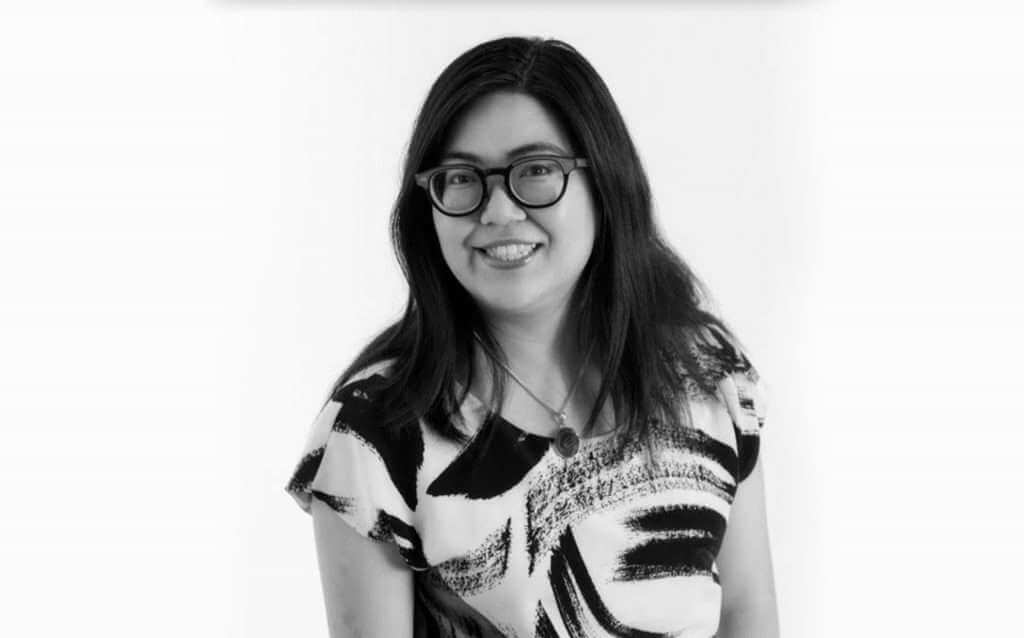 A woman with long dark hair, wearing glasses and a patterned blouse, smiles at the camera in this black-and-white photo, reminiscent of an elegant TouchDesigner creation.