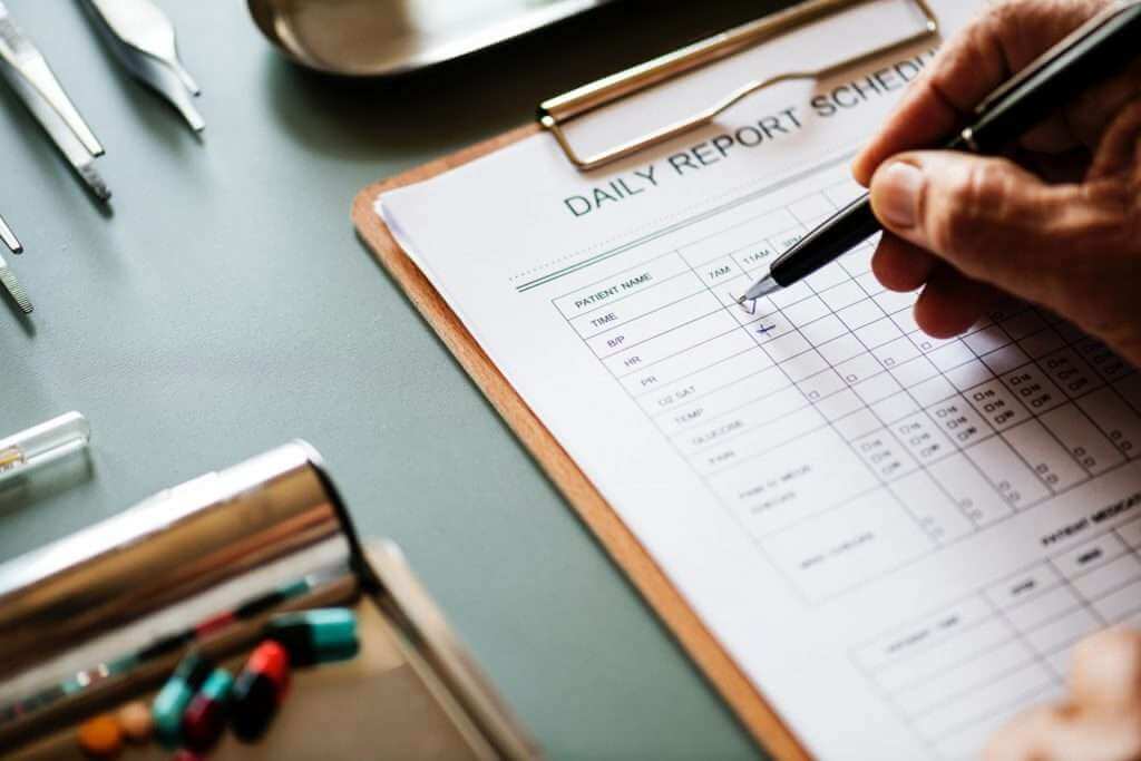 Hand holding pen filling out a "Daily Report Schedule" form on a clipboard. Clipboard sits on a desk with pills, a metal tray, and various medical tools. The scene has the meticulous precision of an artist using TouchDesigner to craft intricate, data-driven visuals.