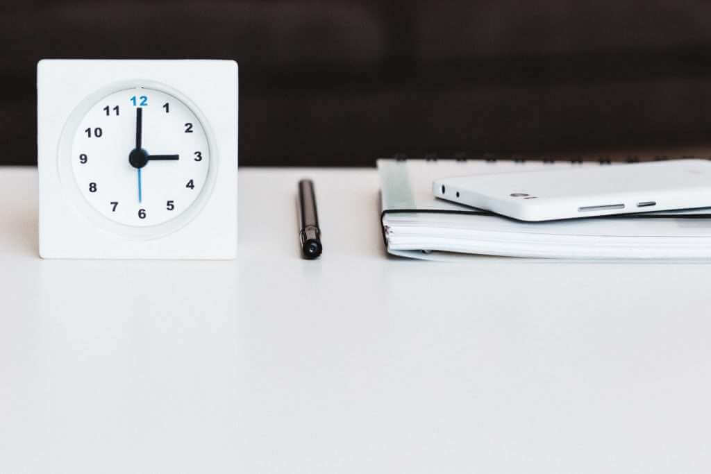 Close-up of a white clock showing 10:10, next to a black pen, a stack of notebooks, and a white smartphone, all placed on a pristine surface that looks straight out of a TouchDesigner setup.