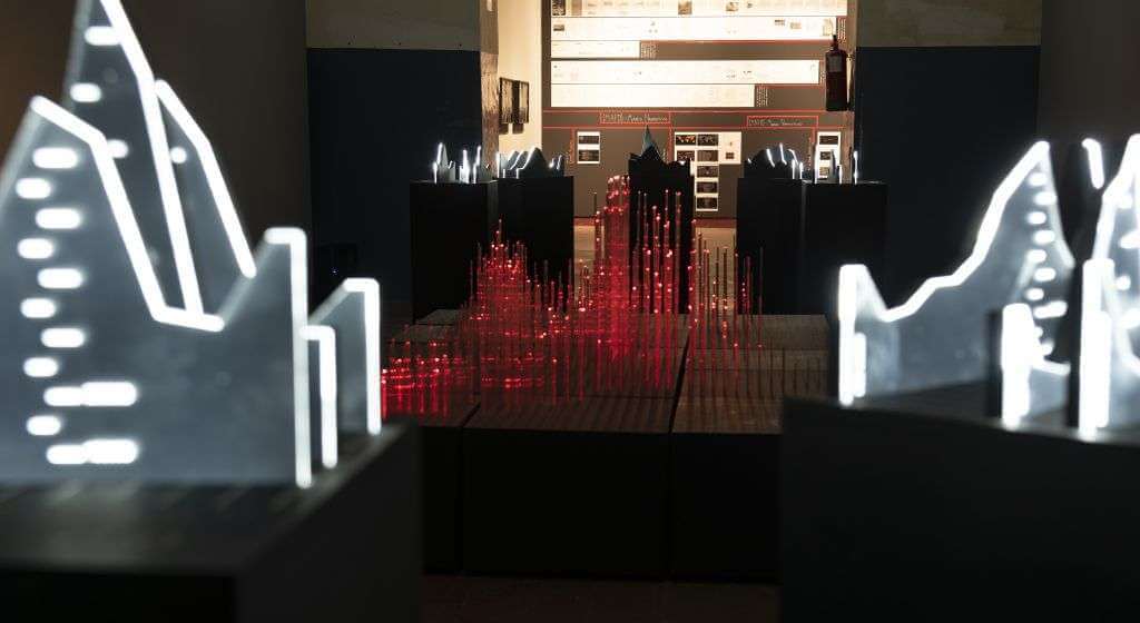 A dark room illuminated by TouchDesigner-created abstract models of cityscapes or statistical graphs, some glowing in red and others in white, displayed on pedestals. The walls are adorned with luminous charts and information panels, creating a mesmerizing digital art experience.