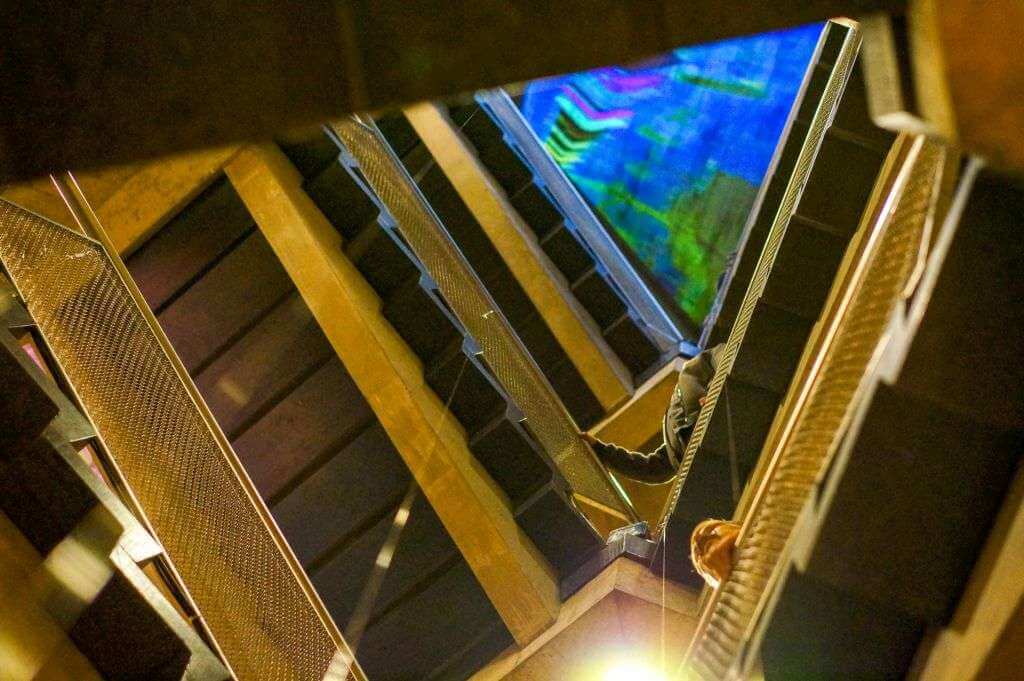 View of a multi-story staircase with metal railings and a colorful stained-glass skylight above, reminiscent of a TouchDesigner visualization. One person is partially visible ascending the stairs.