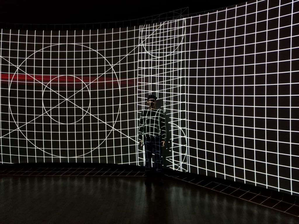 A person stands against a curved wall displaying a black and white grid pattern with geometric designs projected on it, all crafted meticulously using TouchDesigner.