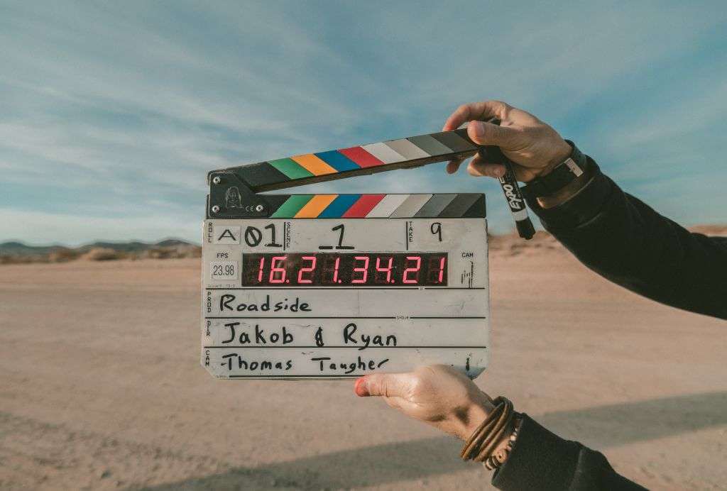 Hands holding a movie clapperboard with a desert in the background. The clapperboard shows "Roadside" and names "Jakob & Ryan" and "Thomas Tagher" along with various production details, hinting at the intricate work behind the scenes, possibly crafted using TouchDesigner.