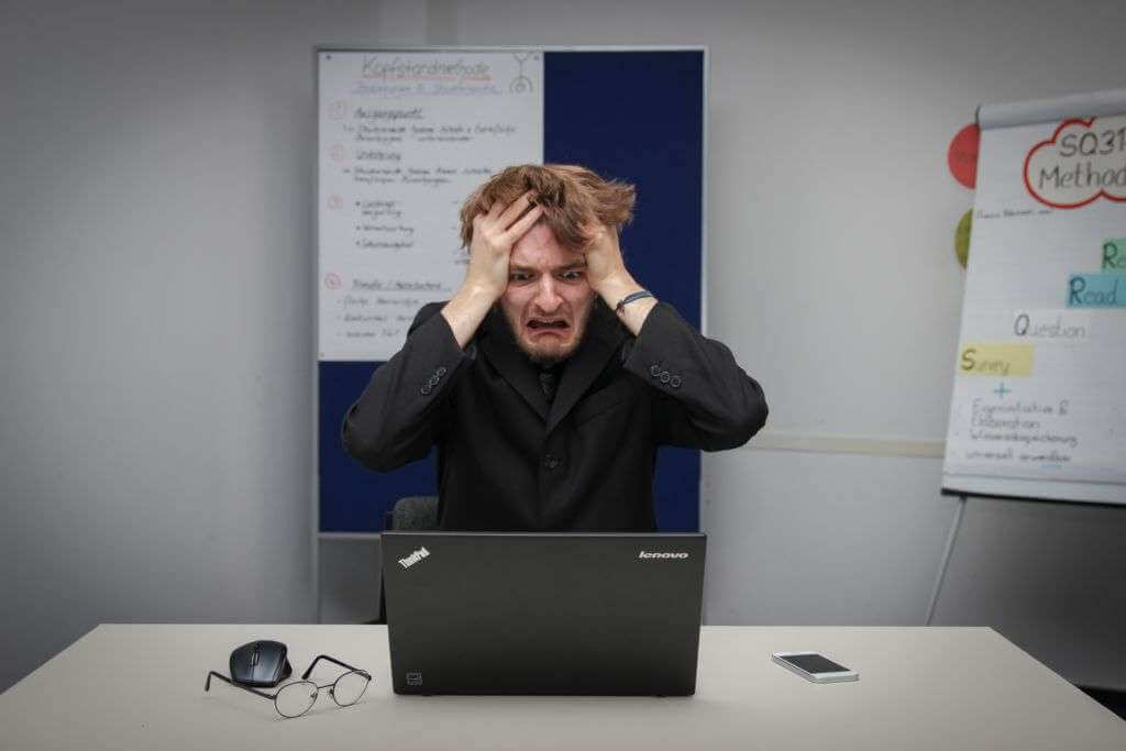 A man sits at a desk with a laptop, holding his head in frustration. A mouse, glasses, and a smartphone are scattered on the desk. Behind him, papers with writing and a colorful flip chart add to the chaos. He seems overwhelmed by his work with TouchDesigner software.