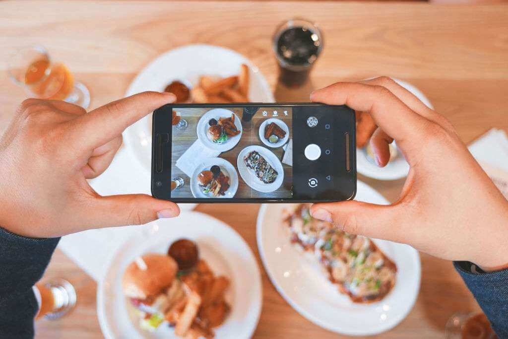 A person using a smartphone to capture a photo of plates with various foods on a table set with several dishes and beverages, almost as if crafting a TouchDesigner masterpiece.