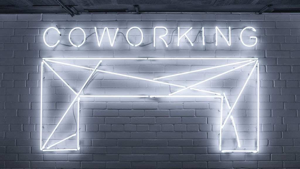 A neon sign on a brick wall displays the word "COWORKING" above a minimalist geometric design resembling a desk, crafted using TouchDesigner for an extra touch of digital artistry.