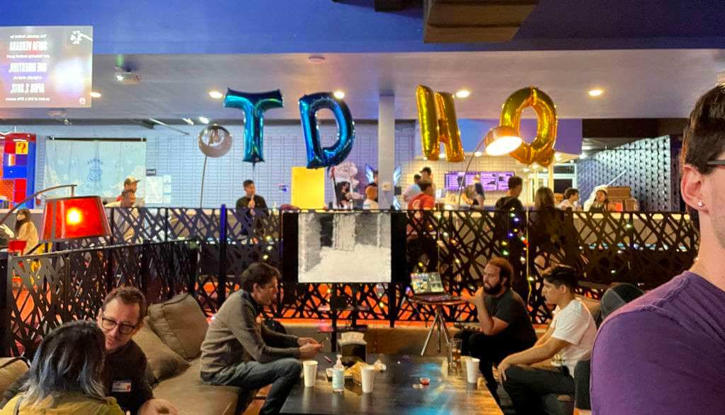 A group of people sit and stand around tables inside a lively room decorated with large balloons spelling "TPIQ," as conversations flow about their latest touchdesigner projects.